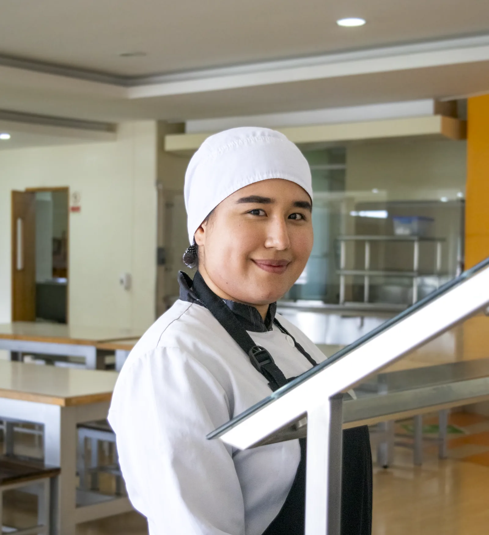 Compañera de cocina con chaqueta y gorro blancos, junto a la línea de servicio de una cafetería institucional